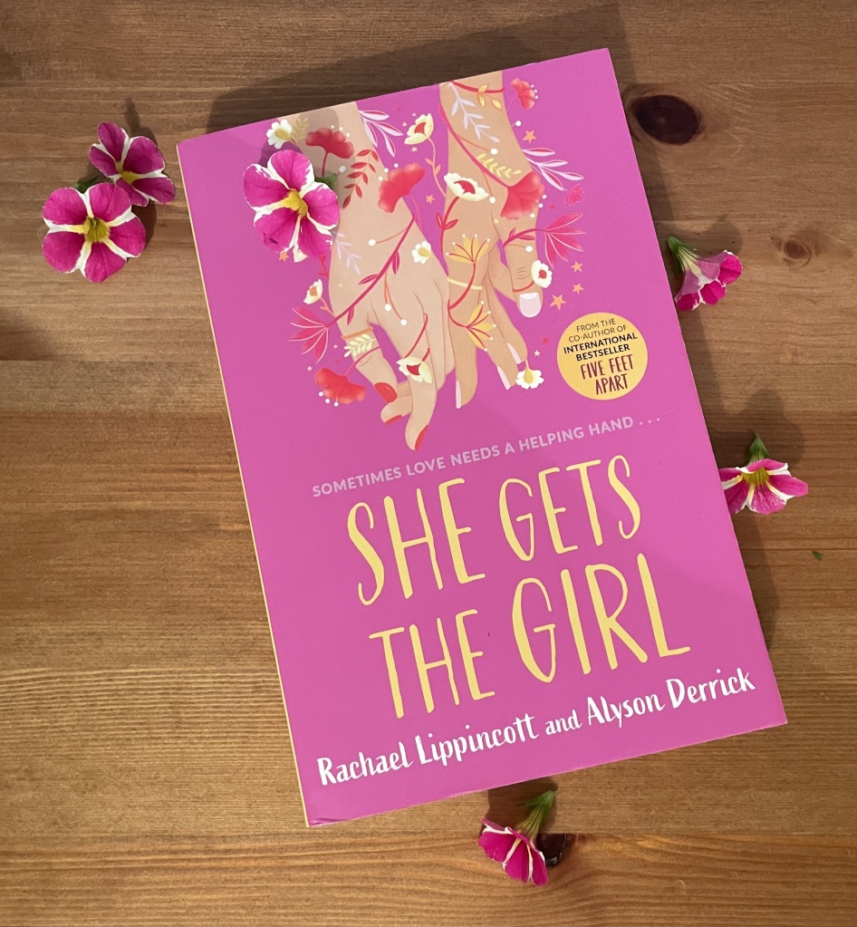 She Gets The Girl paperback against a wooden backdrop surrounded by pink and white flowers.
